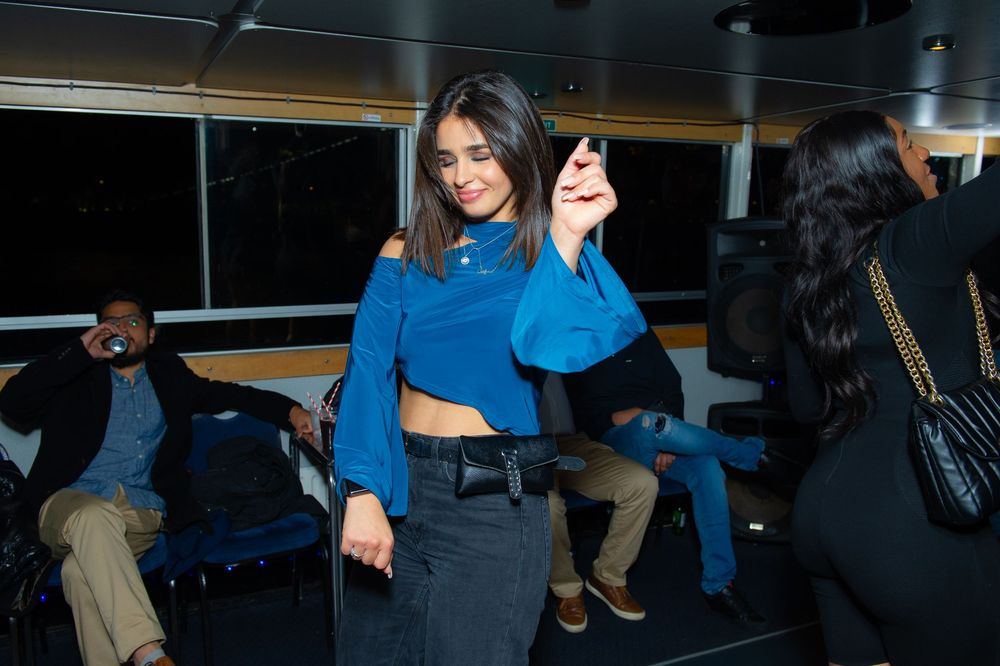 Woman in blue crop top and black skirt dancing at an indoor party event with guests seated in background.