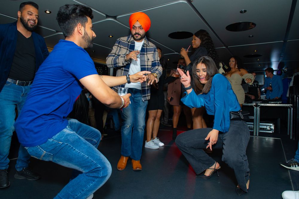 People dancing and celebrating at an indoor party, with one man wearing an orange turban and guests in blue outfits.