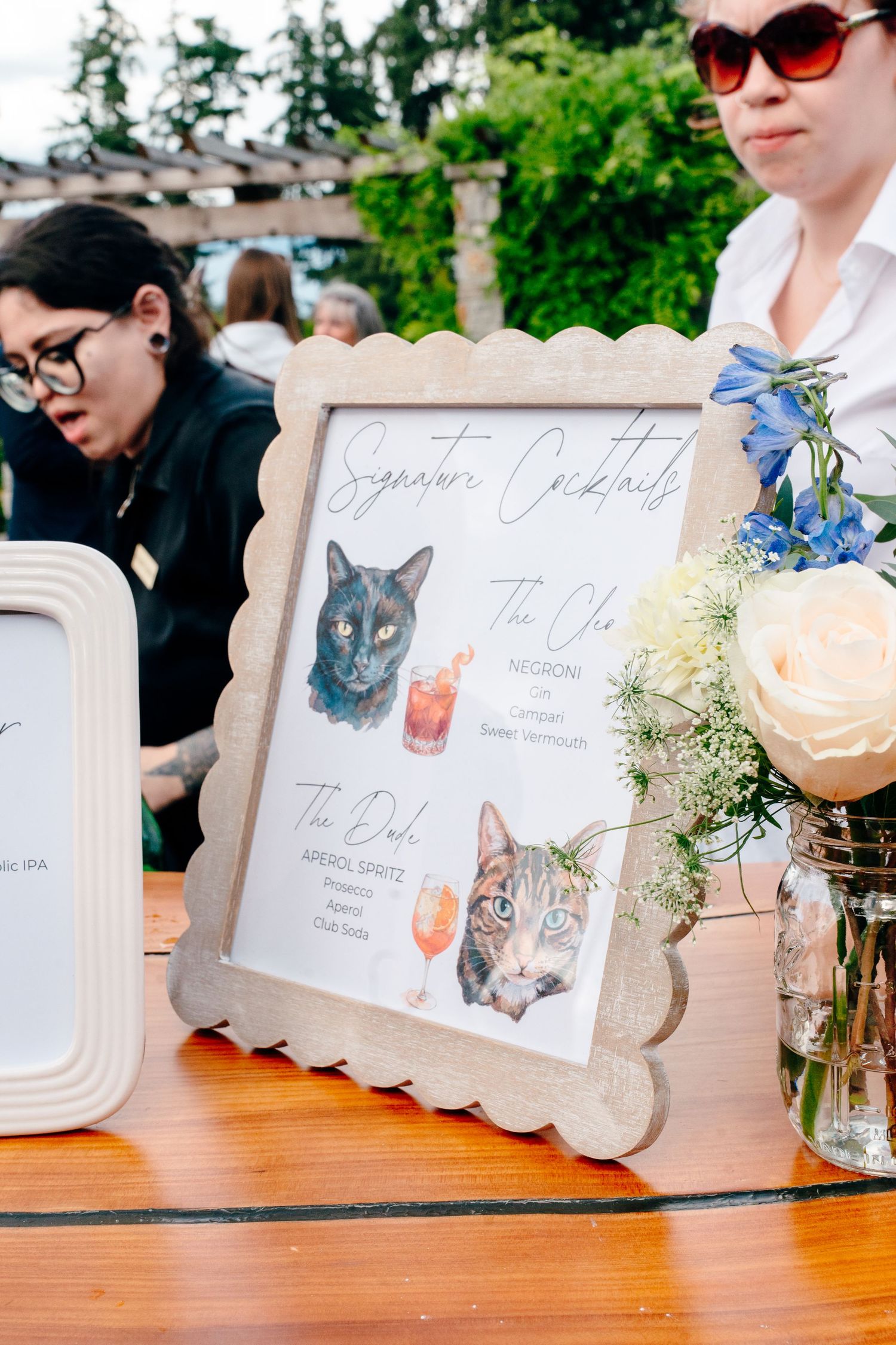 Framed signature cocktails menu with watercolor cat illustrations displayed on a wooden table at an outdoor wedding reception.