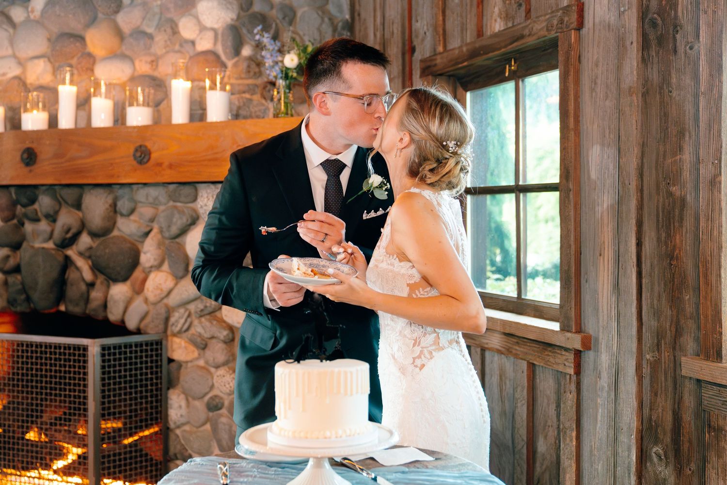 Bride and groom sharing a kiss while cutting their white wedding cake beside a stone fireplace.