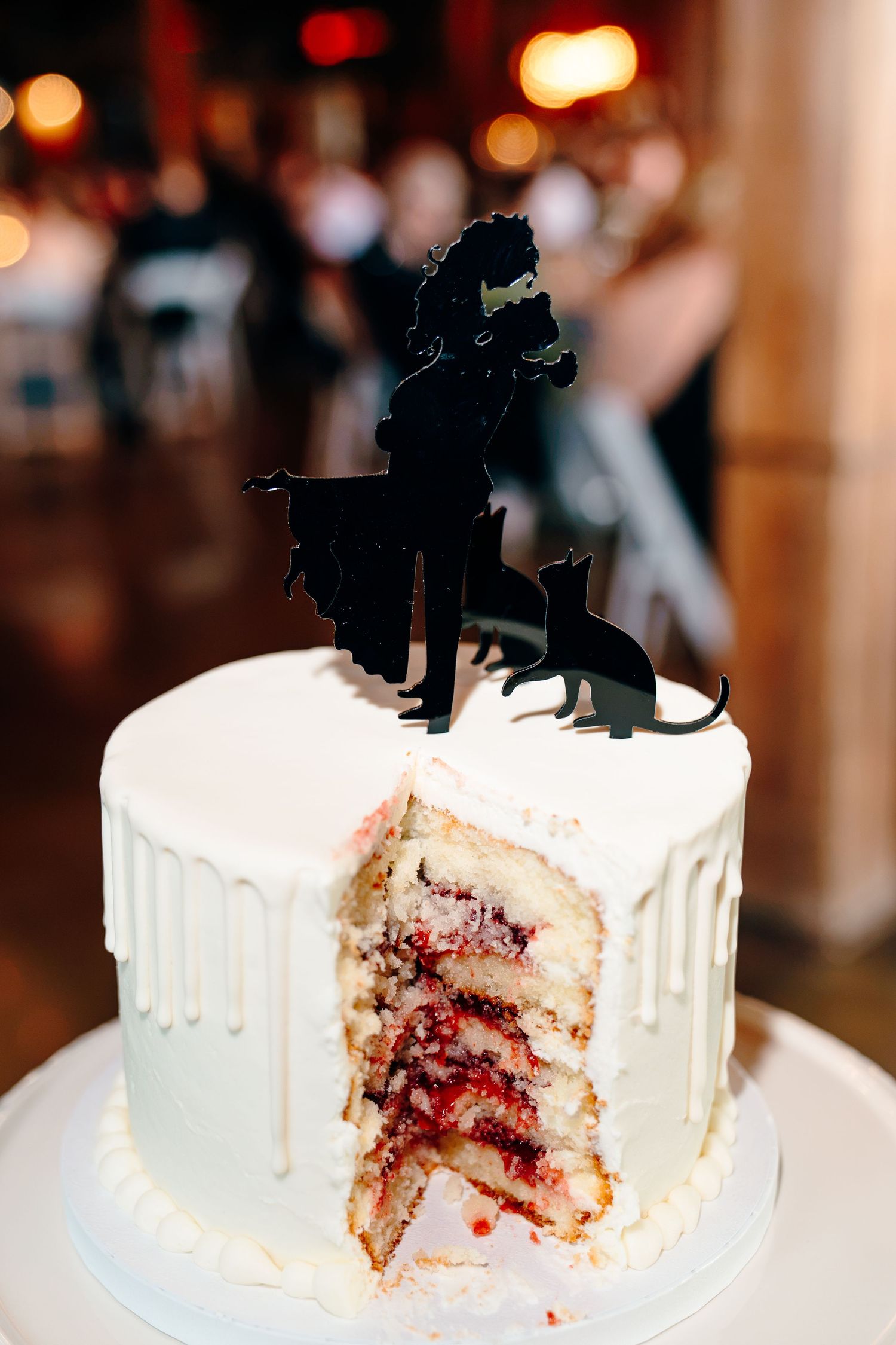 White wedding cake with berry filling, drip frosting, and black silhouette couple with cats topper, slice removed.