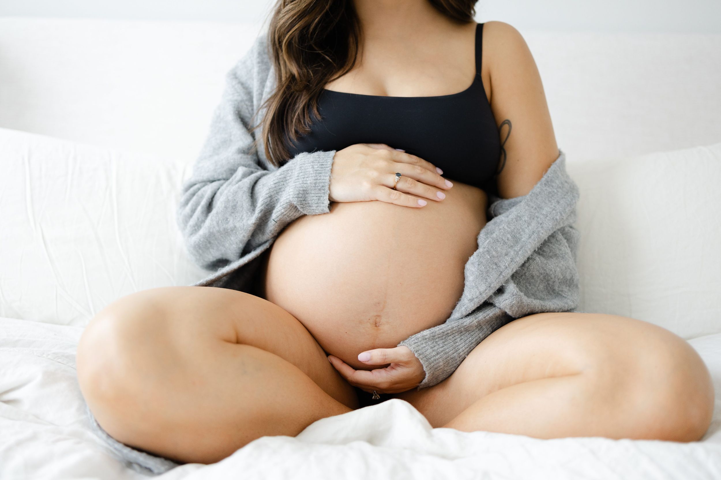 Pregnant woman in black crop top and gray cardigan sitting cross-legged on bed, cradling her bare baby bump.
