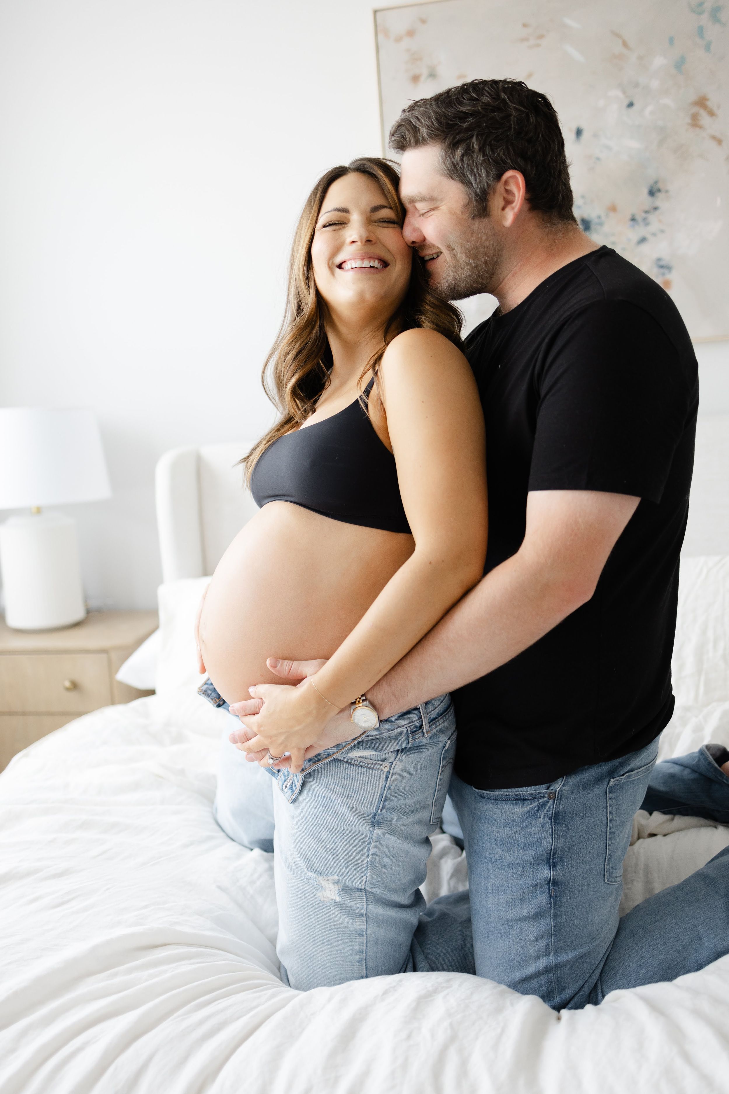 Couple sharing a tender moment on bed, pregnant woman in black bralette and jeans cradling baby bump with partner kissing her cheek.