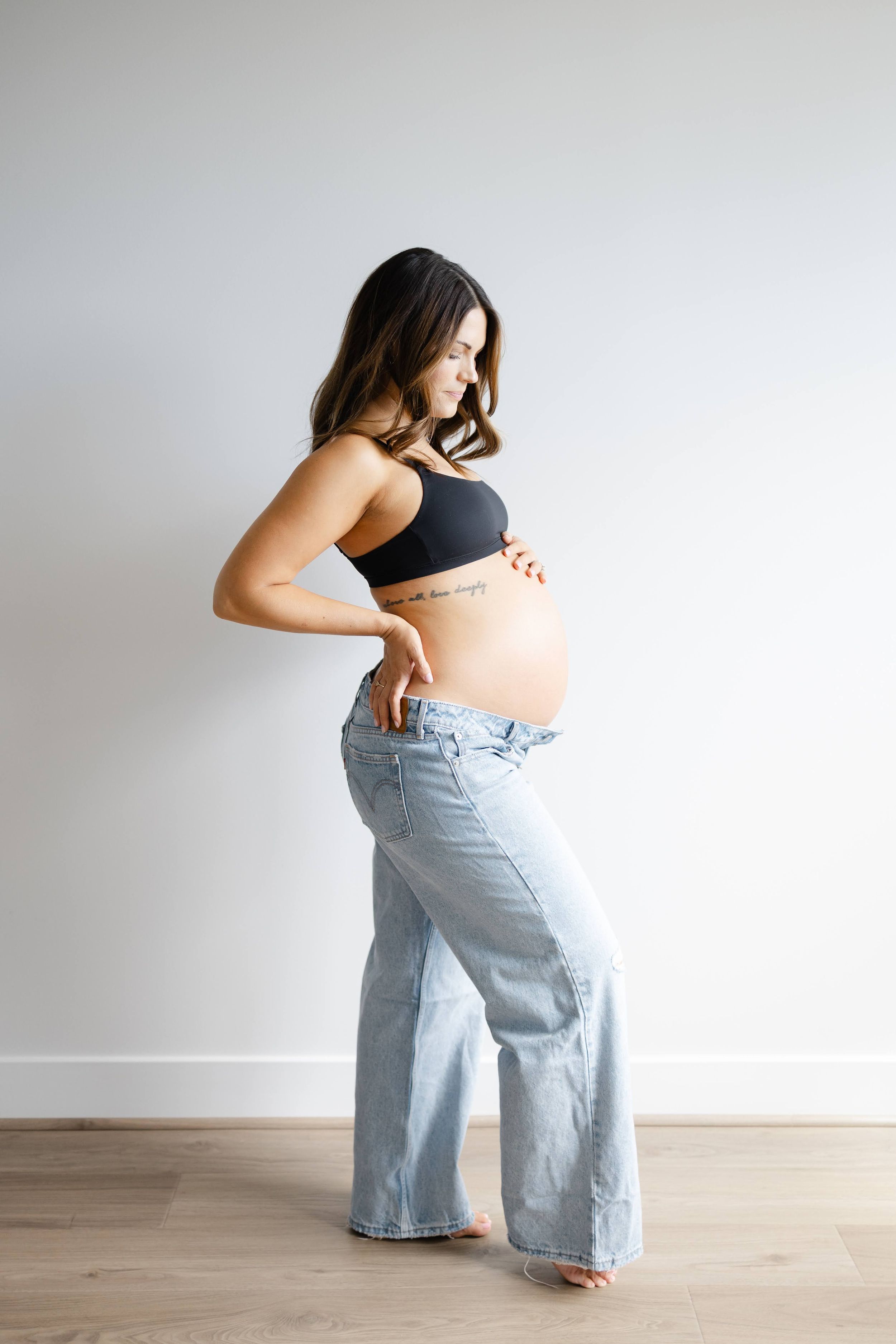 Pregnant woman in black crop top and wide-leg jeans cradling baby bump, standing against white wall.