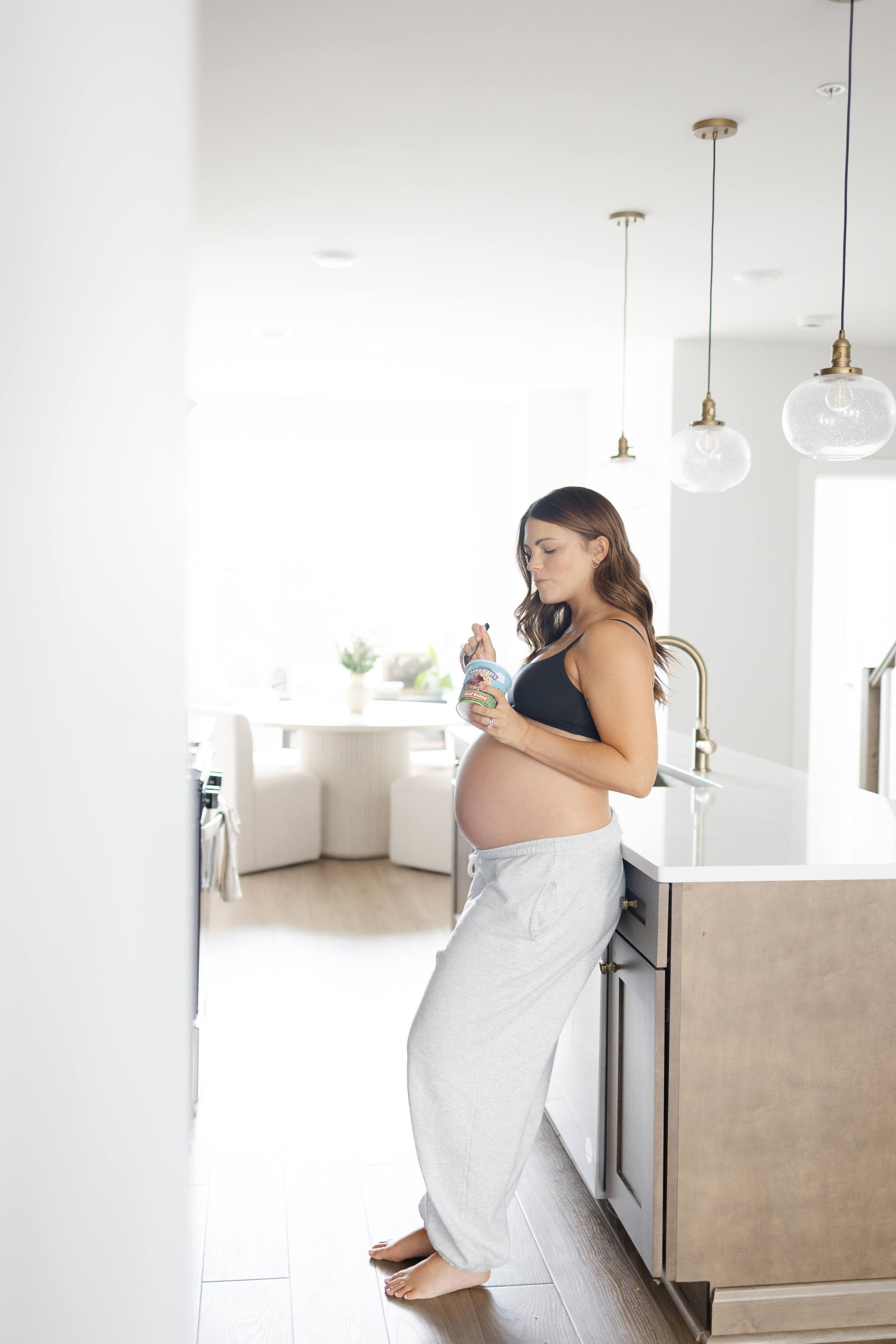 Pregnant woman in black crop top and white pants standing in modern kitchen holding a baby item.