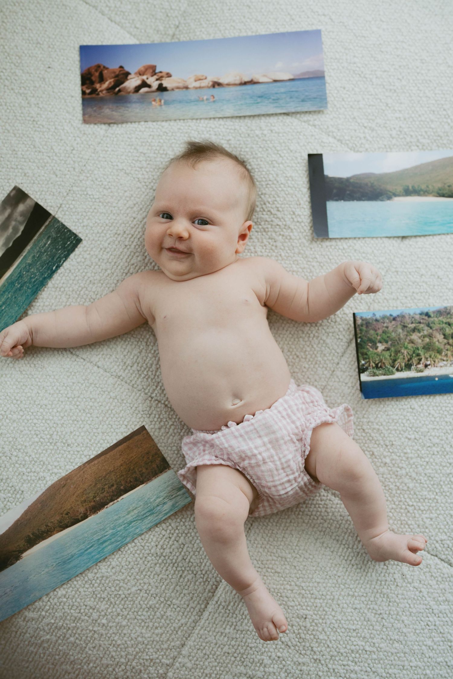 Smiling baby in pink bloomers lying on a cream carpet surrounded by colorful tropical beach photo prints.