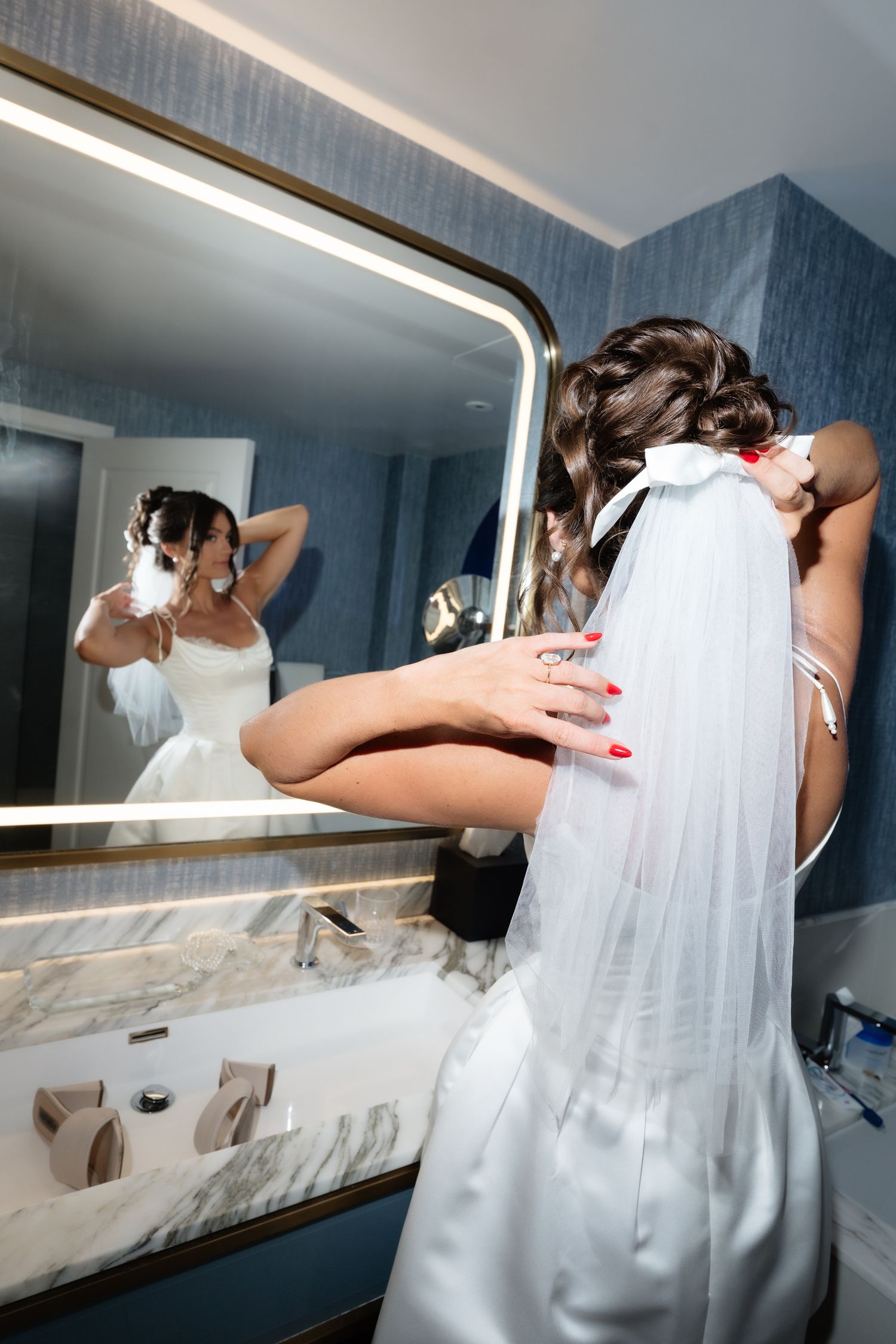 Bride in white wedding dress and veil adjusts hair while looking in illuminated bathroom mirror.
