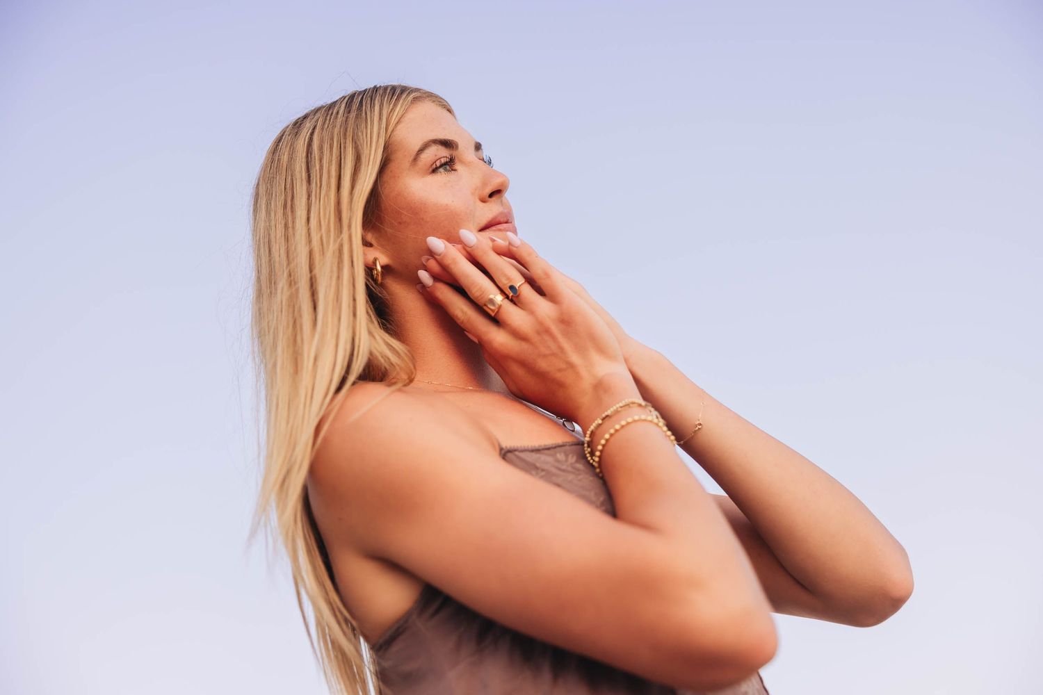 Blonde woman with gold bracelet touching her face against a clear blue sky in a serene outdoor portrait.