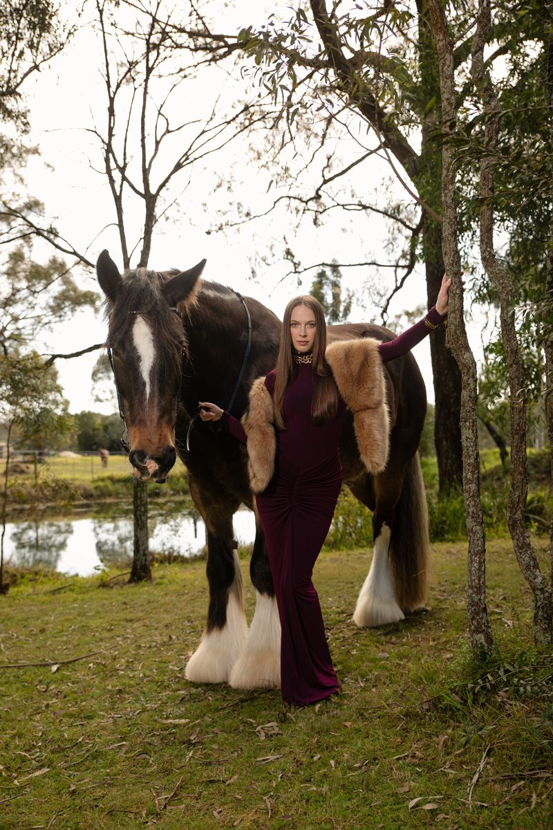 Woman in burgundy gown with fur stole stands beside a shire horse near trees and a pond during a photoshoot in sydney