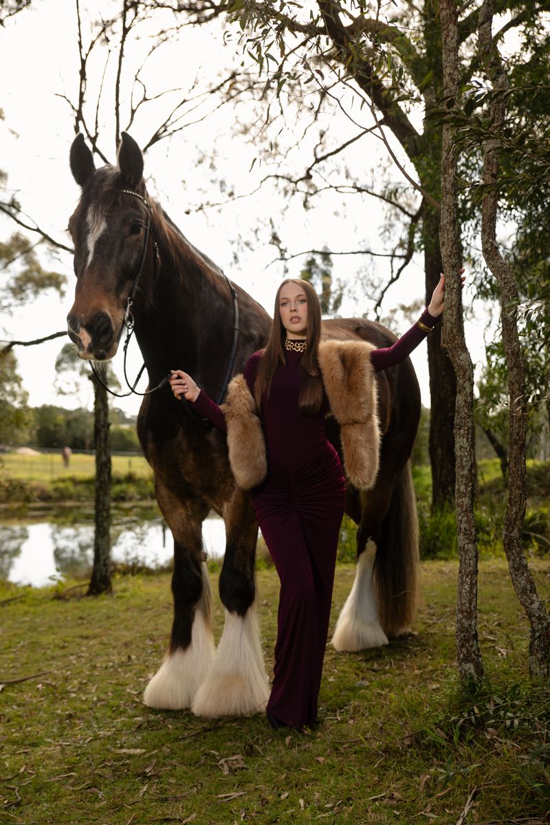 Woman in burgundy gown with fur stole poses beside a brown shire horse near trees and a pond photoshoot with a horse in sydney