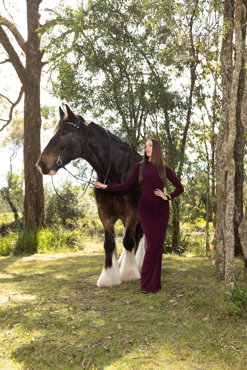 woman in burgundy dress poses with a large shire horse in a sunlit forest setting in sydney photoshoot with a horse