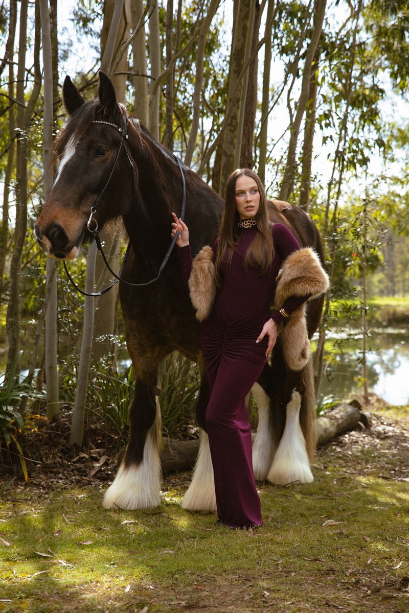 Woman in burgundy outfit with fur stole poses beside a large shire horse in a forest setting during a photoshoot in sydney