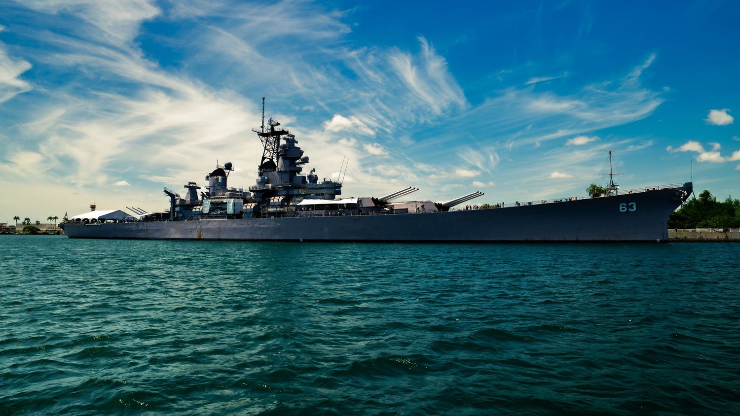 The gray battleship USS Missouri, hull number 63, sits moored at Pearl Harbor under a blue sky with wispy clouds in Honolulu, Hawaii.