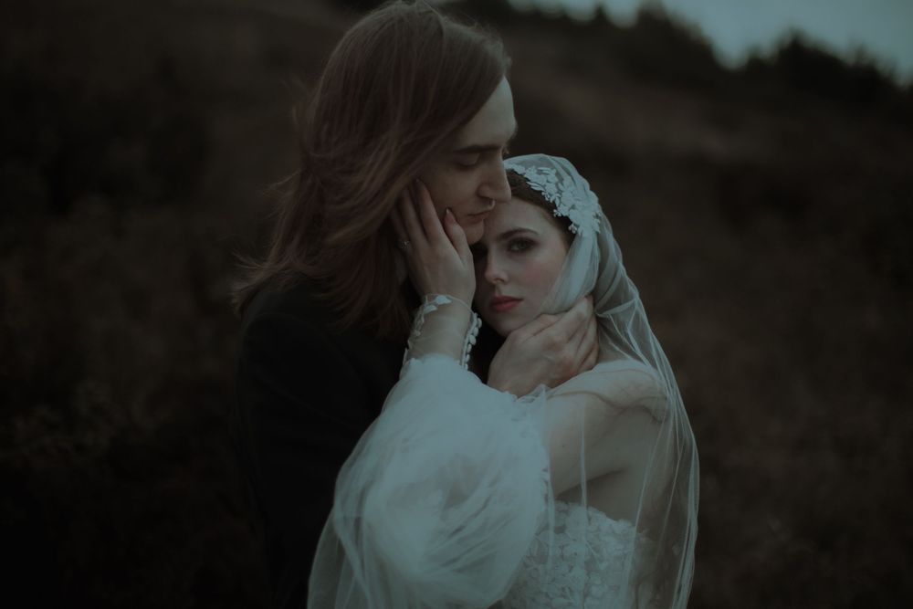 A bride in a white lace veil and gown shares an intimate moment with her partner outdoors at dusk.