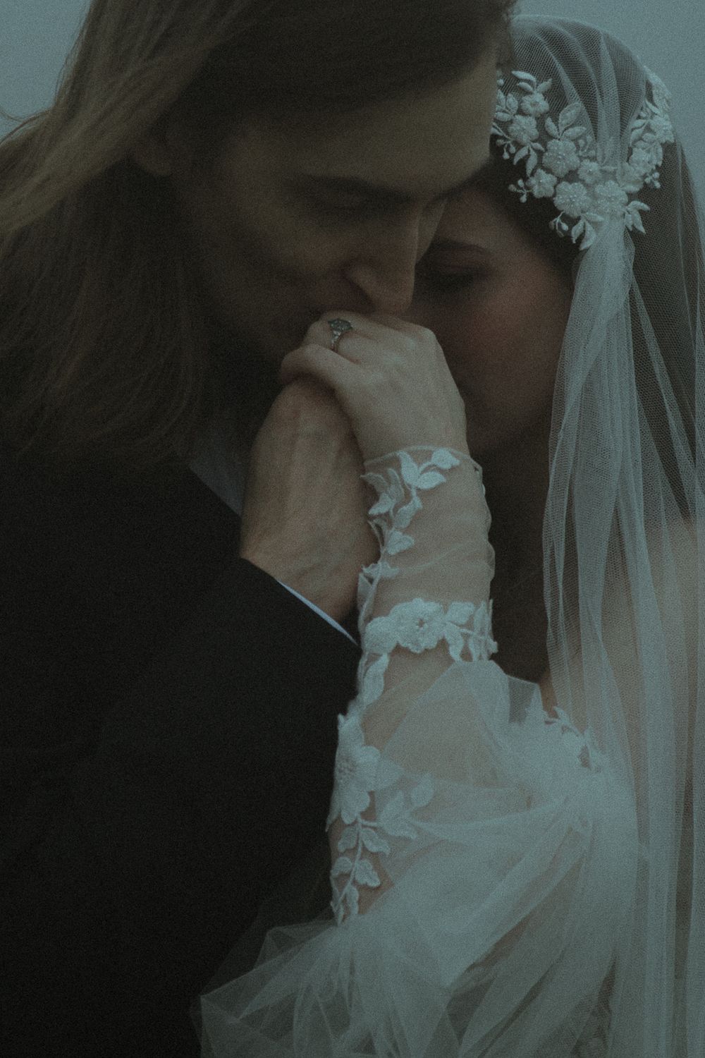 Groom in black suit kissing bride's hand, bride wearing lace wedding dress and floral veil headpiece.