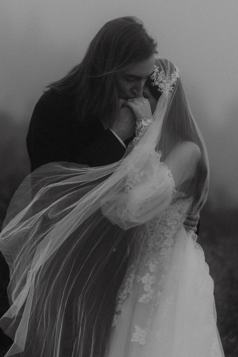 Black and white photo of bride and groom embracing, veil flowing in wind.