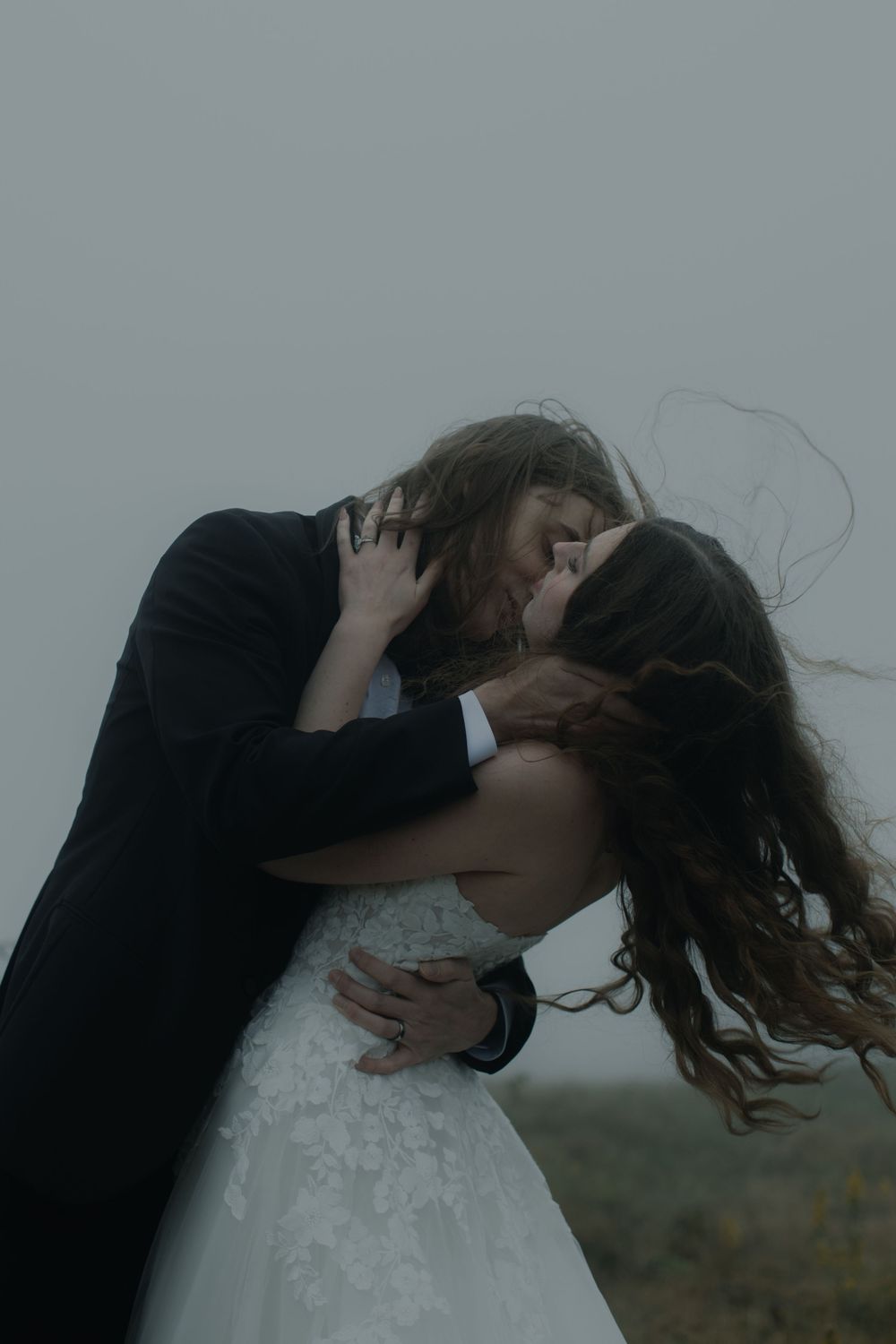A bride and groom share a passionate kiss in the wind, dressed in white lace gown and black suit on a moody day.