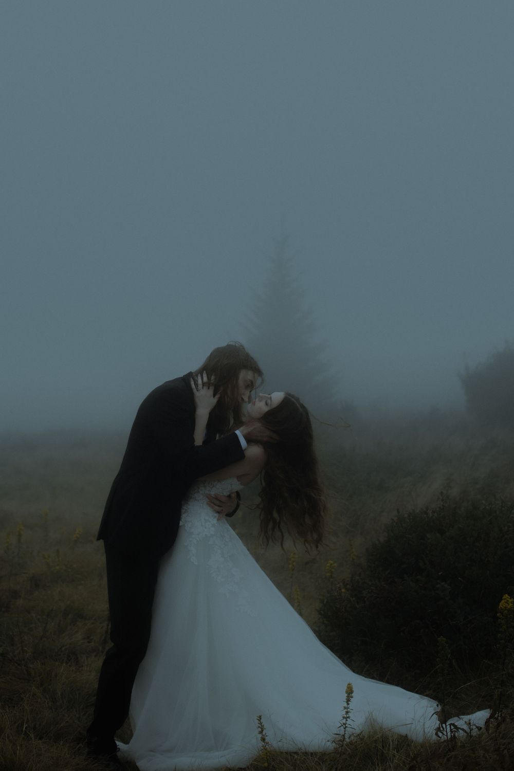 Bride and groom sharing romantic kiss in misty foggy outdoor wedding setting.