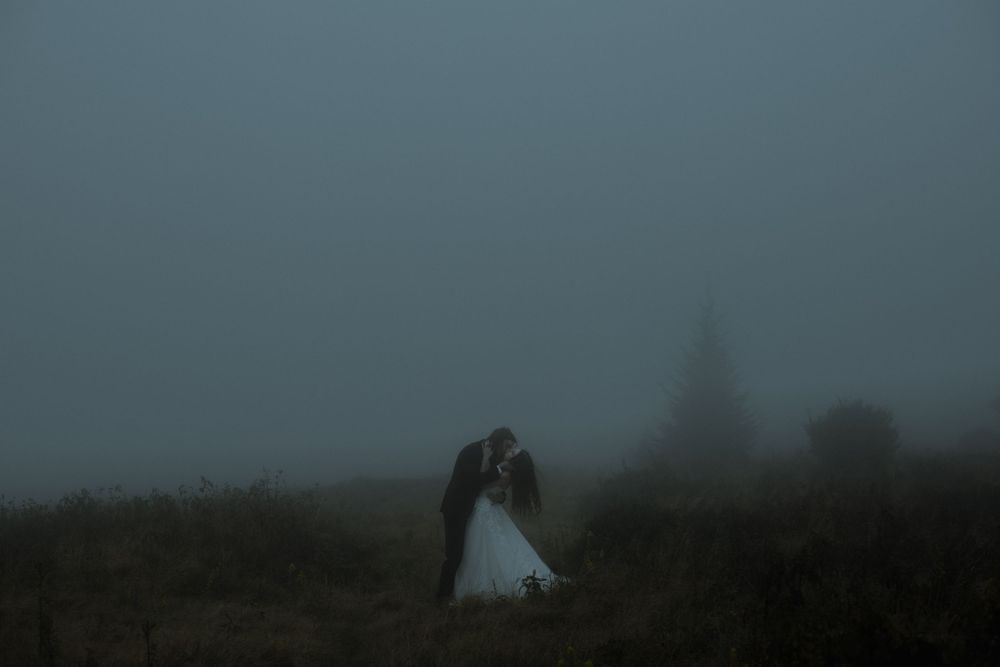 Couple sharing a romantic kiss in a misty foggy field, bride in white gown, moody dark atmospheric wedding photo.