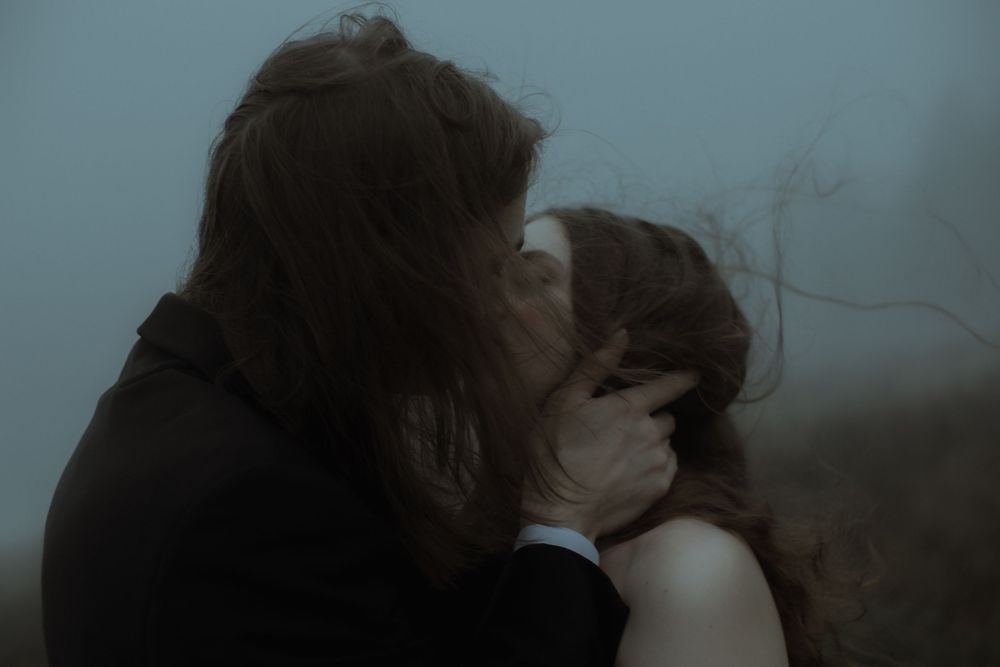 Couple sharing a romantic kiss in moody foggy atmosphere, man in dark suit embracing woman with flowing hair.