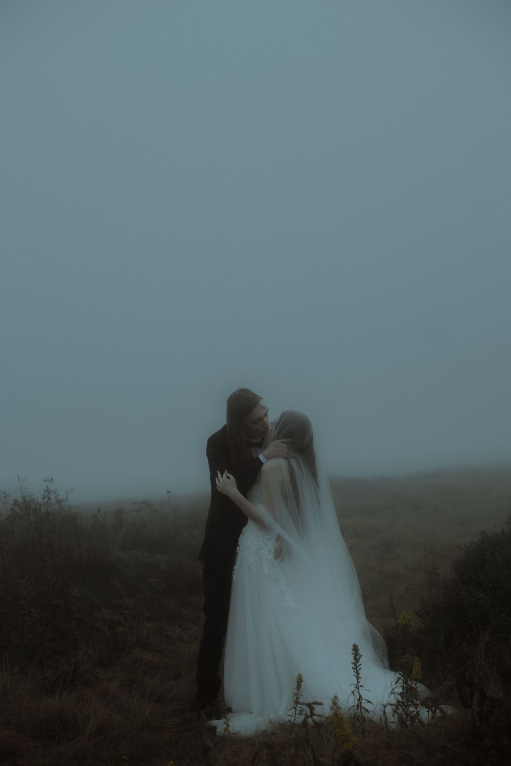 Bride and groom embrace in misty foggy outdoor setting, bride wearing white gown and veil for romantic wedding photo.