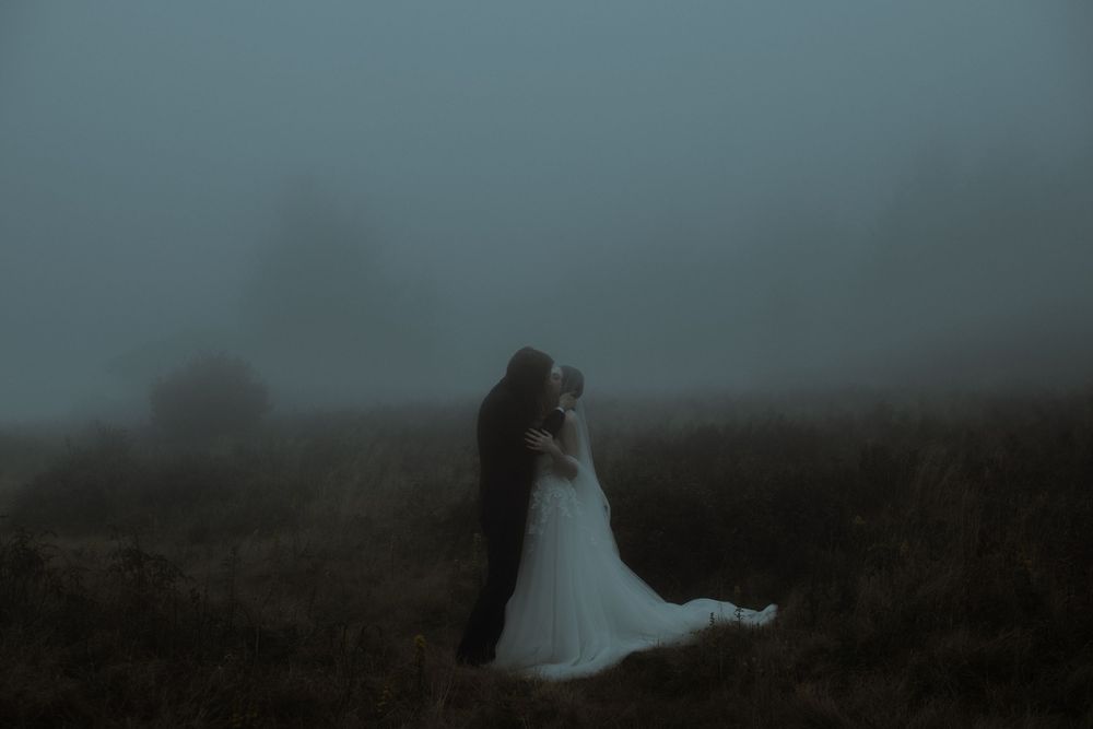 A couple in wedding attire embraces in a misty, foggy field at dusk, creating a moody romantic atmosphere.