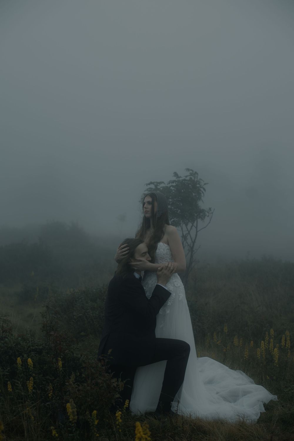 A bride in a white gown embraces a groom in a black suit amid a misty, foggy field with wildflowers.