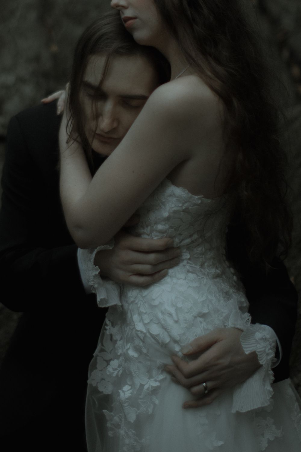 A bride in a white lace gown embraces a man in a dark suit in a moody, romantic outdoor setting.