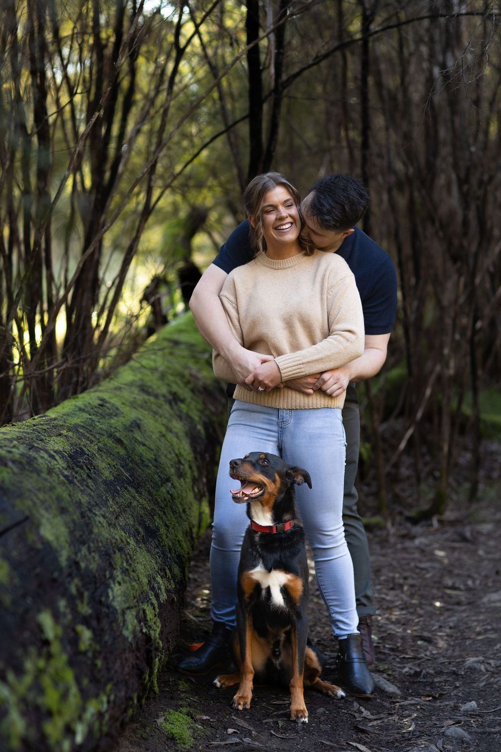 Couple embracing in a forest, smiling, with a black and tan dog sitting in front of a moss-covered log.