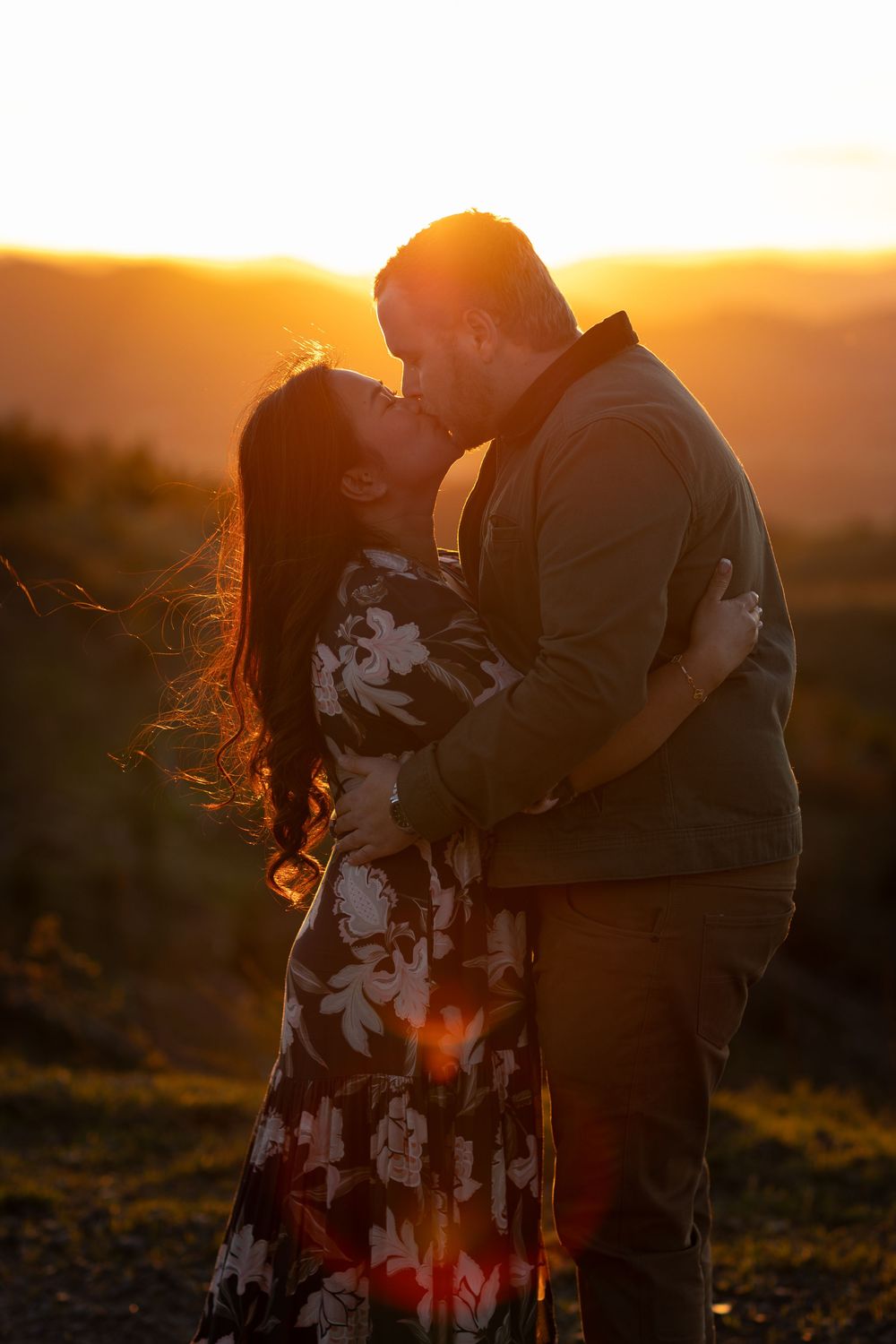 Couple sharing a romantic kiss at golden sunset, woman in floral dress, man in jacket, with warm glowing hills behind them.