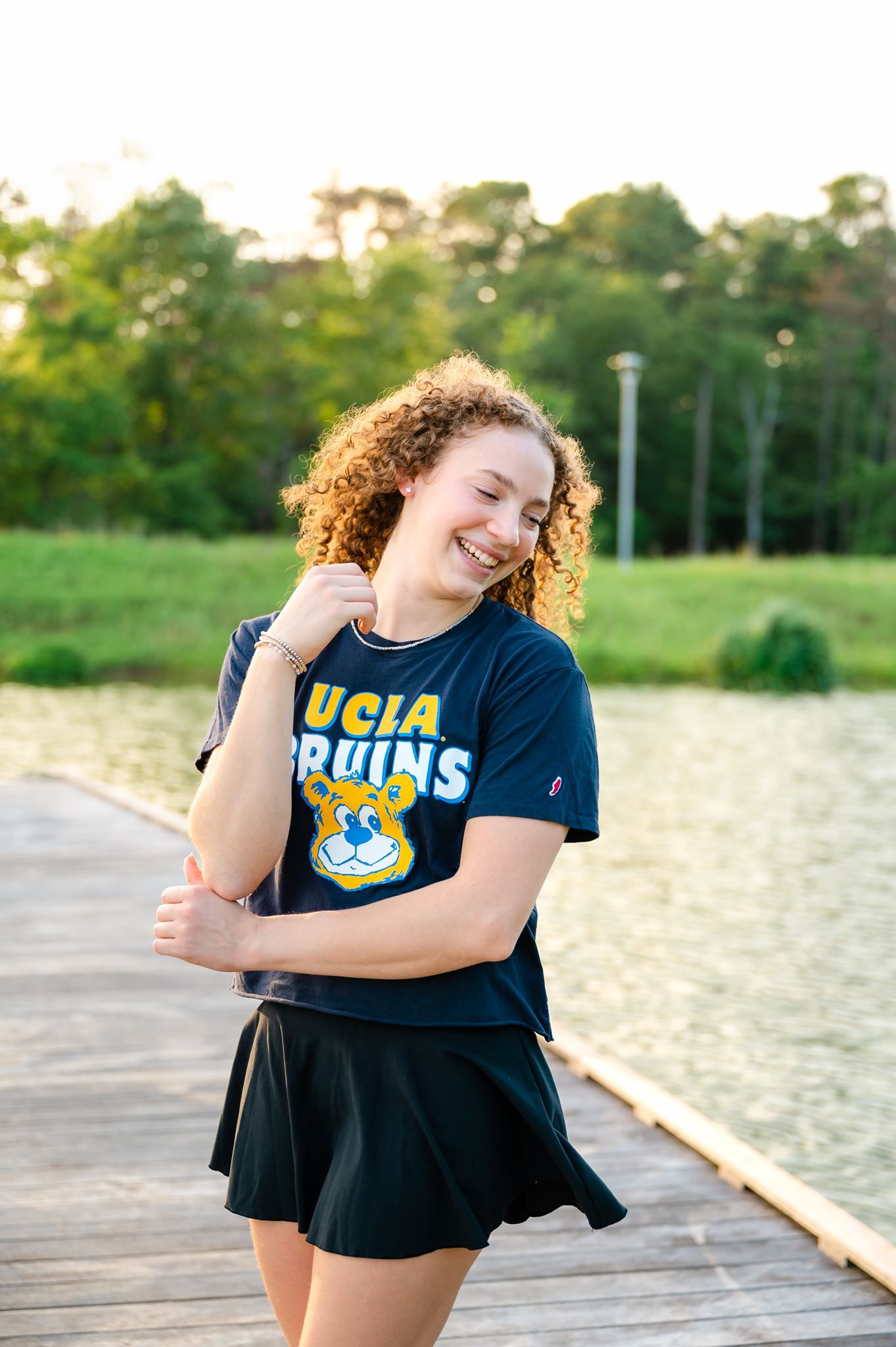 High school senior in UCLA Bruins navy crop tee and black skirt standing on a wooden dock by a lake.