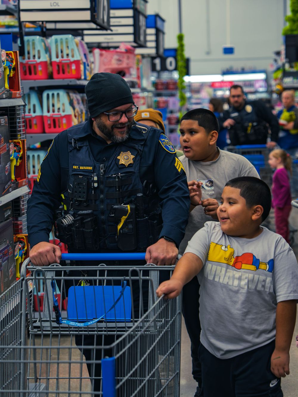 A uniformed police officer pushes a shopping cart down a toy aisle, smiling with two children beside him as they shop together at the event.