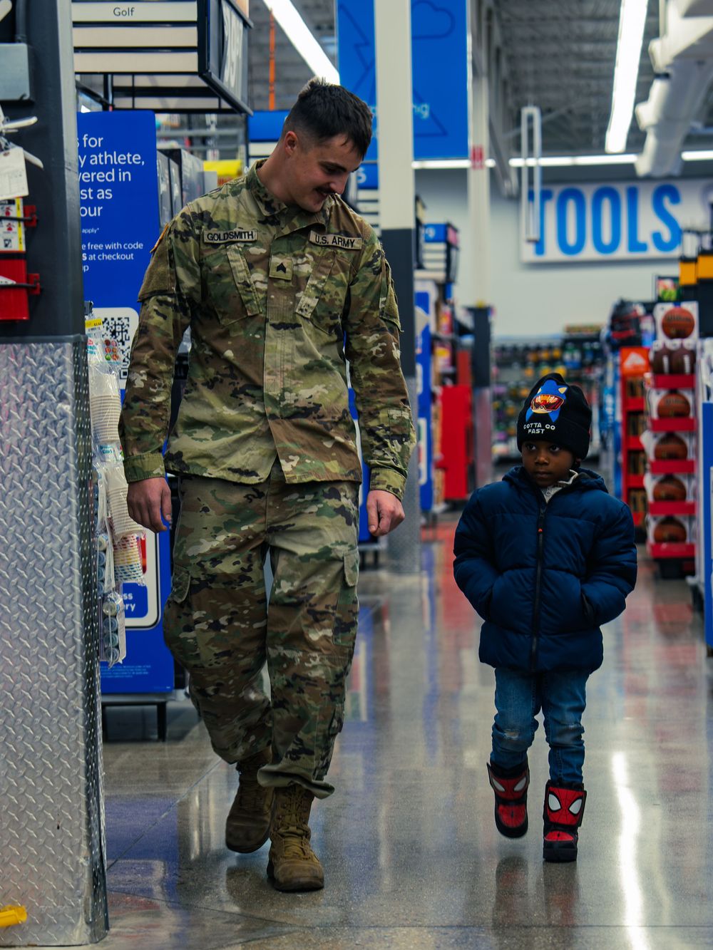 National Guard soldier walks down a store aisle beside a young child in a blue coat and character boots as they shop together at the event.