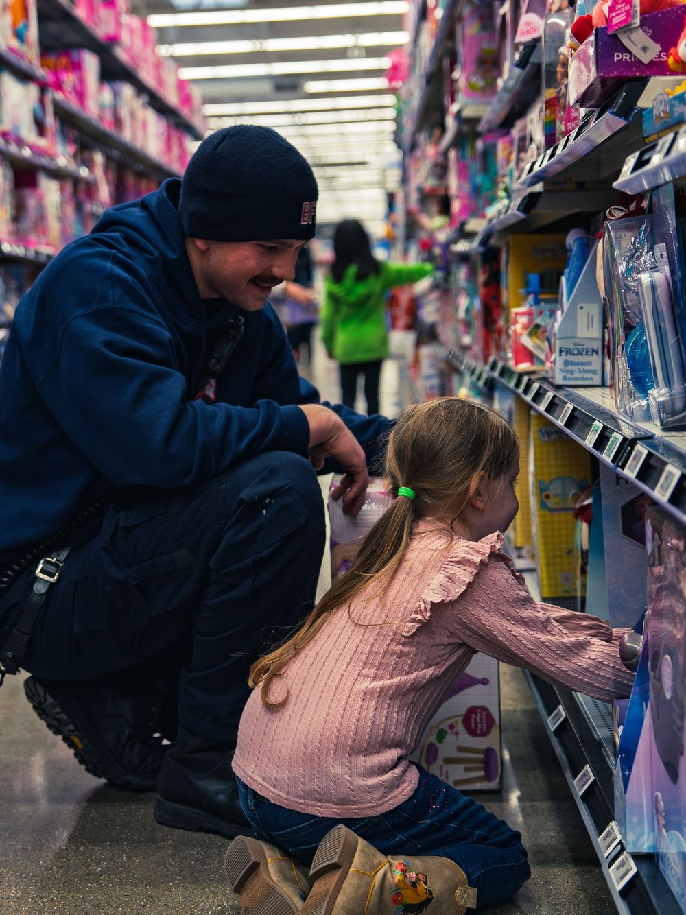 A firefighter in a navy department beanie crouches beside a young child reaching for a toy on the shelf as they shop the toy aisle together.