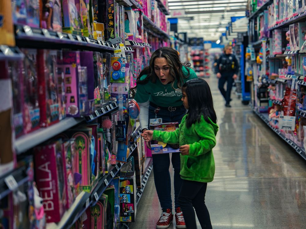 A volunteer in a green shirt helps a young child in a bright green jacket pick a toy from the shelf as an officer walks the aisle behind.