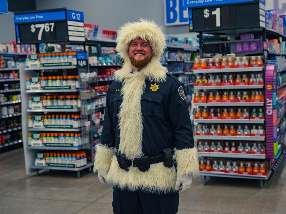 A smiling police officer in a fur-trimmed winter hood and coat over his uniform stands in a store aisle at the holiday shopping event.
