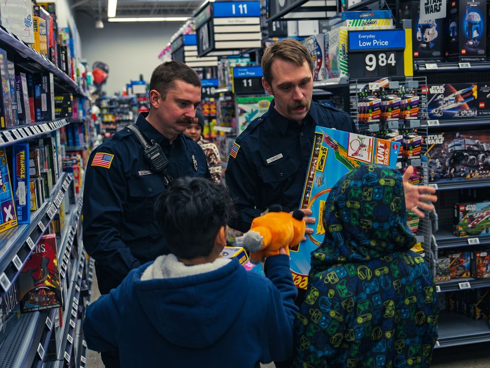 Two firefighters in navy uniforms help two children pick out toys in the games aisle, one holding up a race track set to show them.