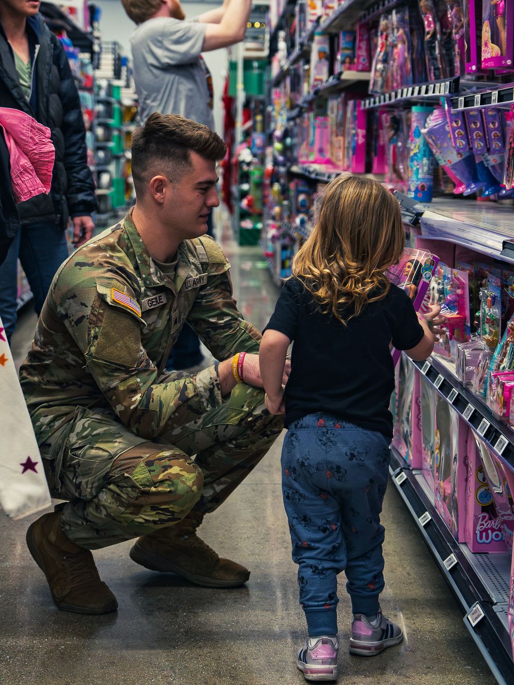 A National Guard soldier kneels down to a young child holding a doll from the shelf as they shop the toy aisle together at the event.