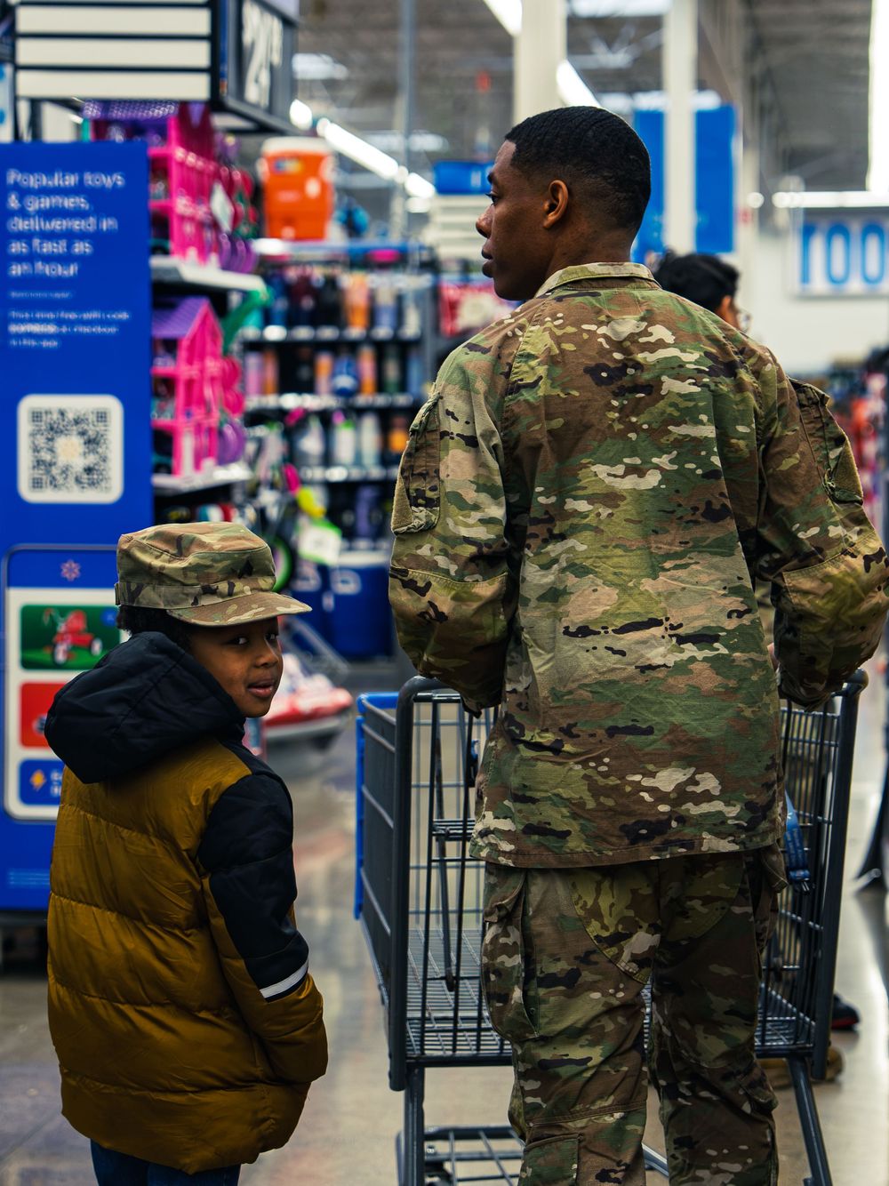 A National Guard soldier pushes a shopping cart down a store aisle beside a young child wearing a camo military cap.