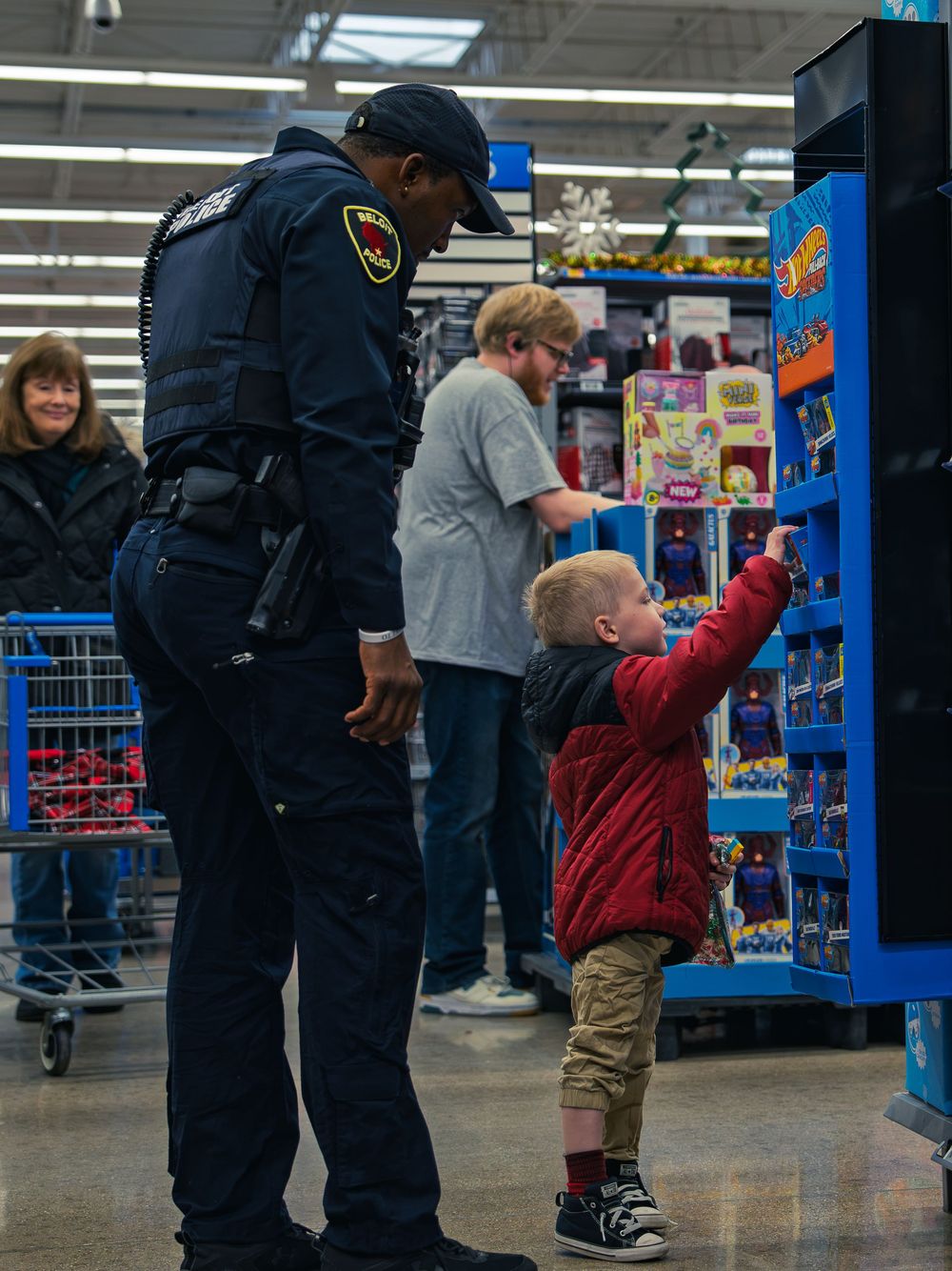 A police officer stands beside a young child in a red jacket who reaches for a toy car on a store display as shoppers browse behind them.