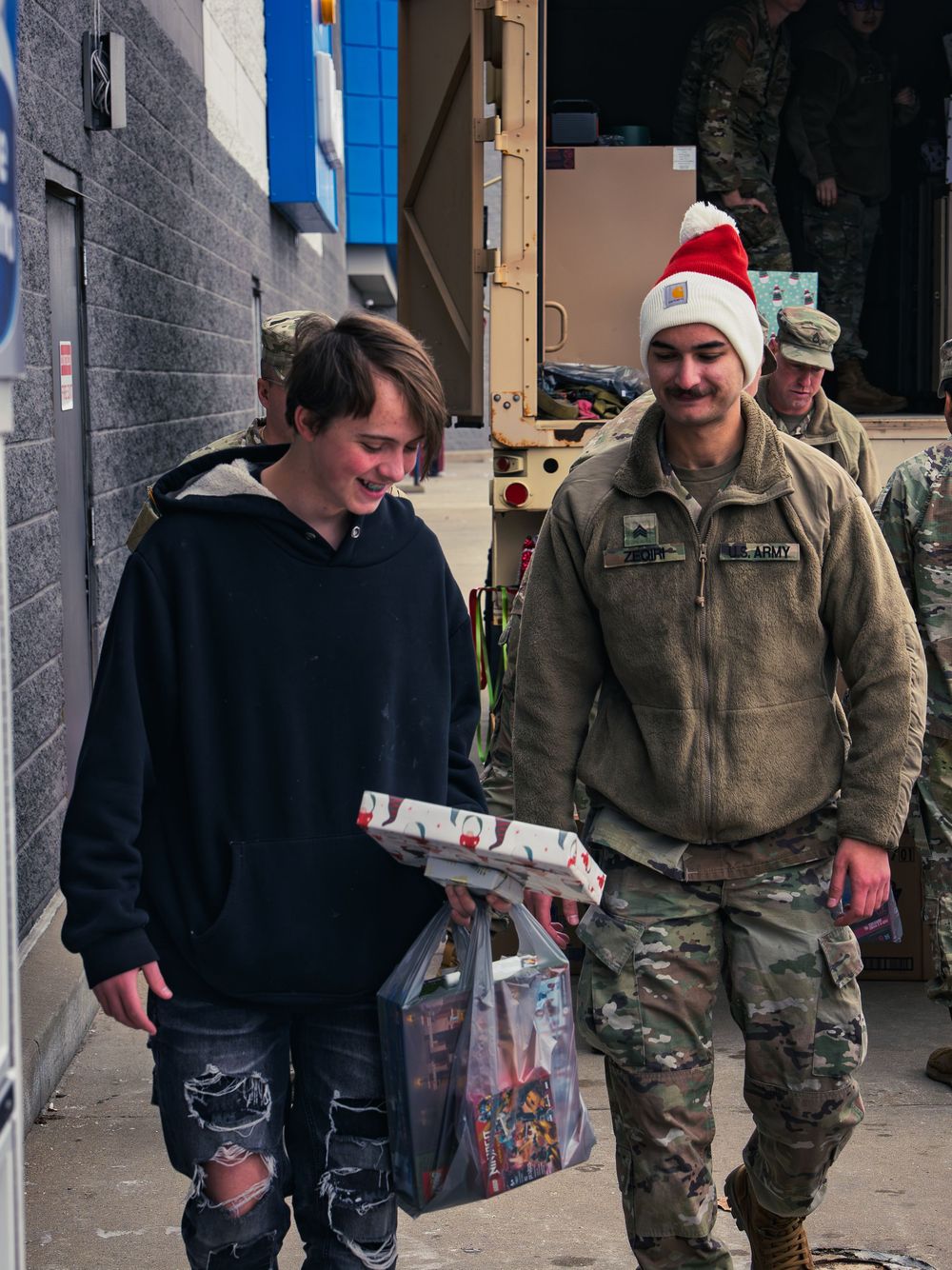 A smiling teen carries a wrapped gift and a bag of toys beside a National Guard soldier in a Santa-style hat as more soldiers unload behind.