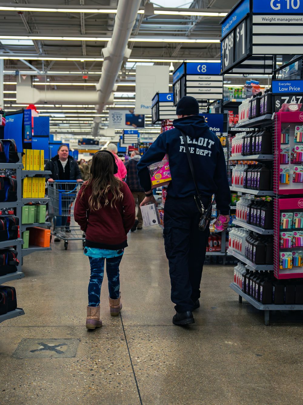 A firefighter carrying toys walks down a store aisle beside a young child, both seen from behind as they shop together at the event.