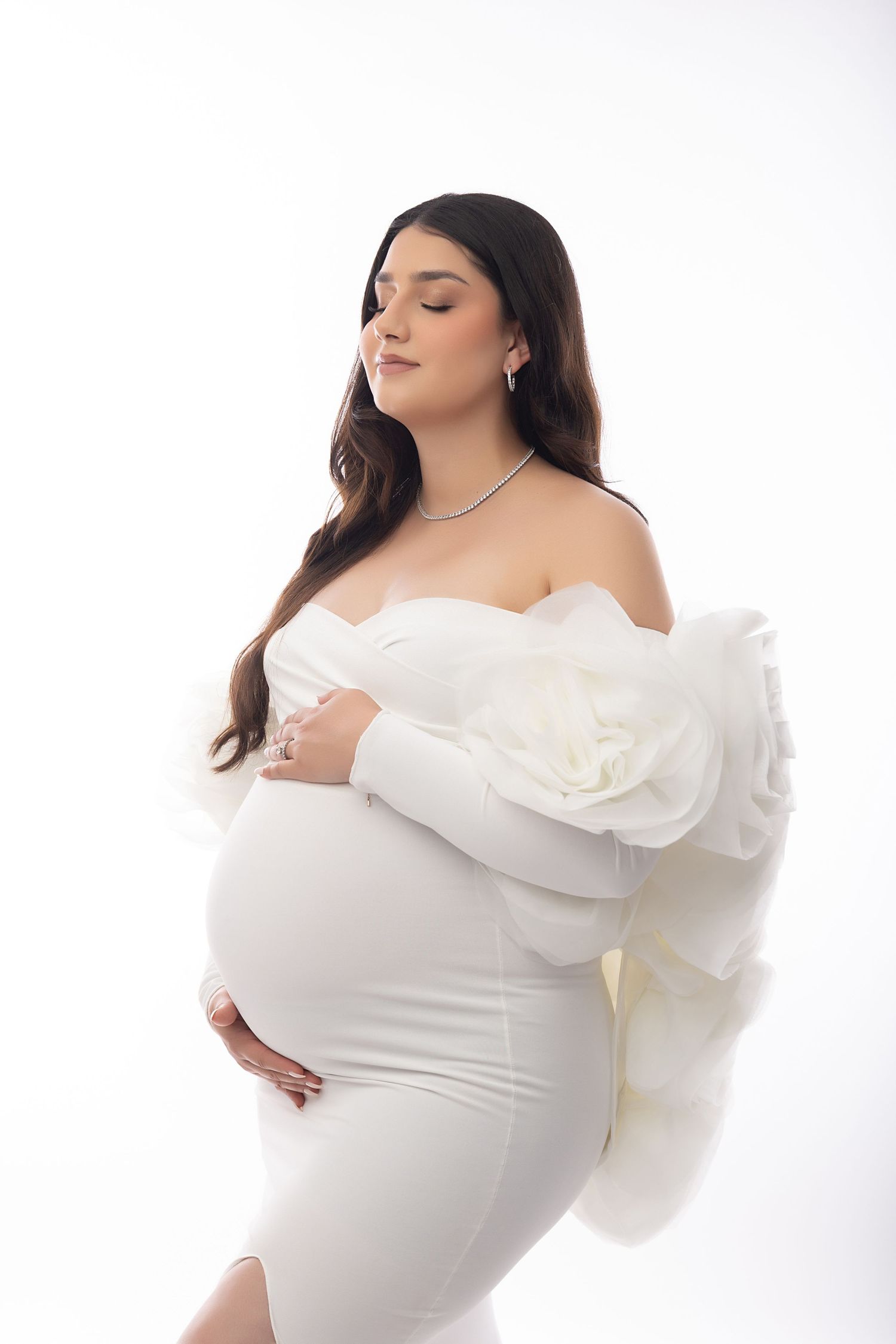 Pregnant woman in white off-shoulder gown with ruffled sleeves, cradling baby bump, eyes closed, on white background.