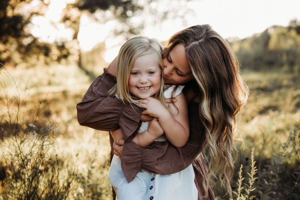 A mother hugging and kissing her smiling young blonde daughter outdoors in a sunlit field.