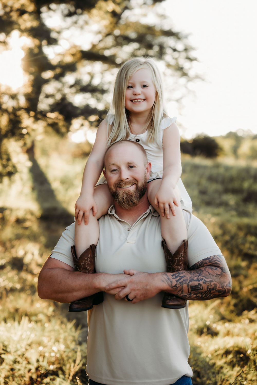 A bald bearded tattooed father carries his smiling blonde daughter on his shoulders outdoors in golden sunlight.