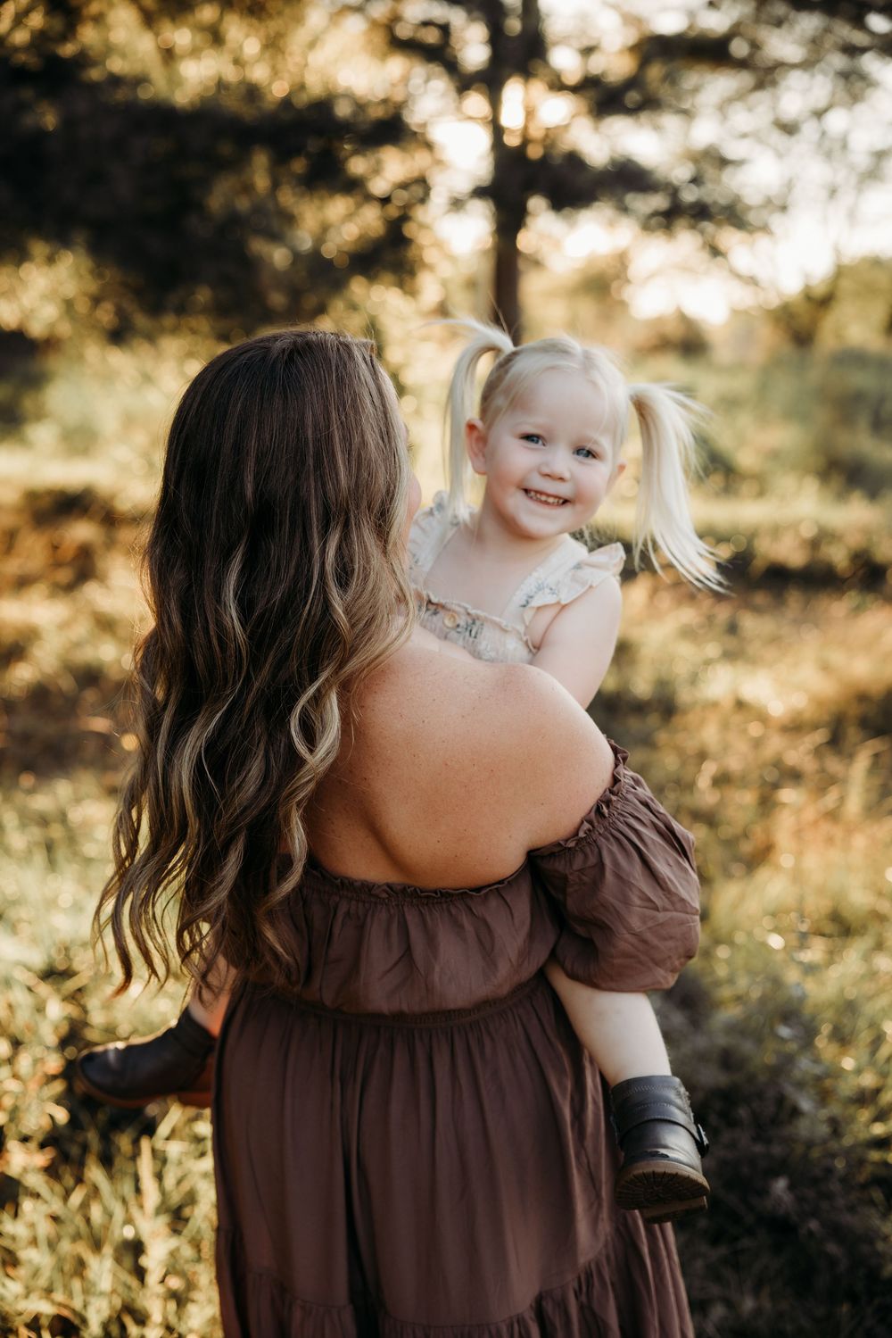 A mother in a brown dress holds her smiling toddler girl with pigtails outdoors in golden sunlight.