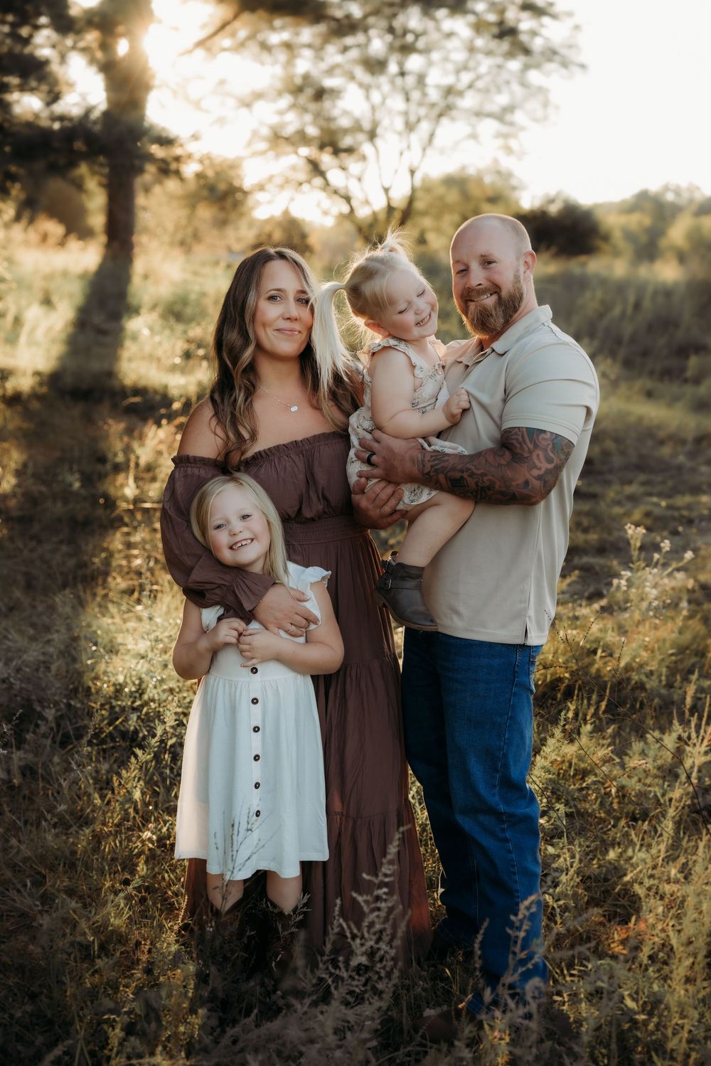 Family of four posing outdoors at golden hour, parents holding two young daughters in a sunlit field.