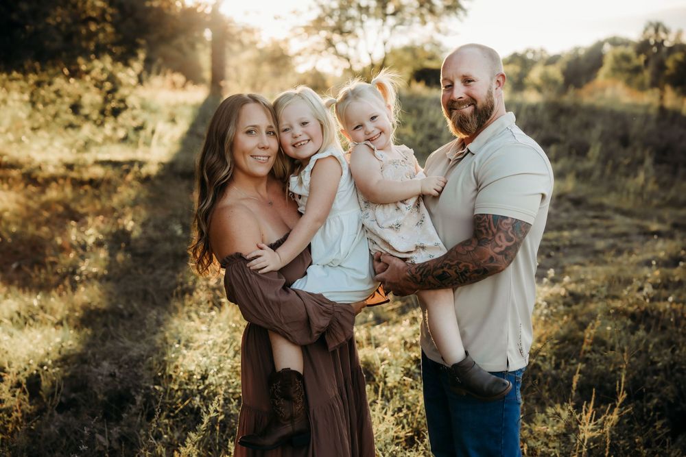 Family of four posing outdoors in golden sunlight, parents holding two young daughters in a sunny meadow.