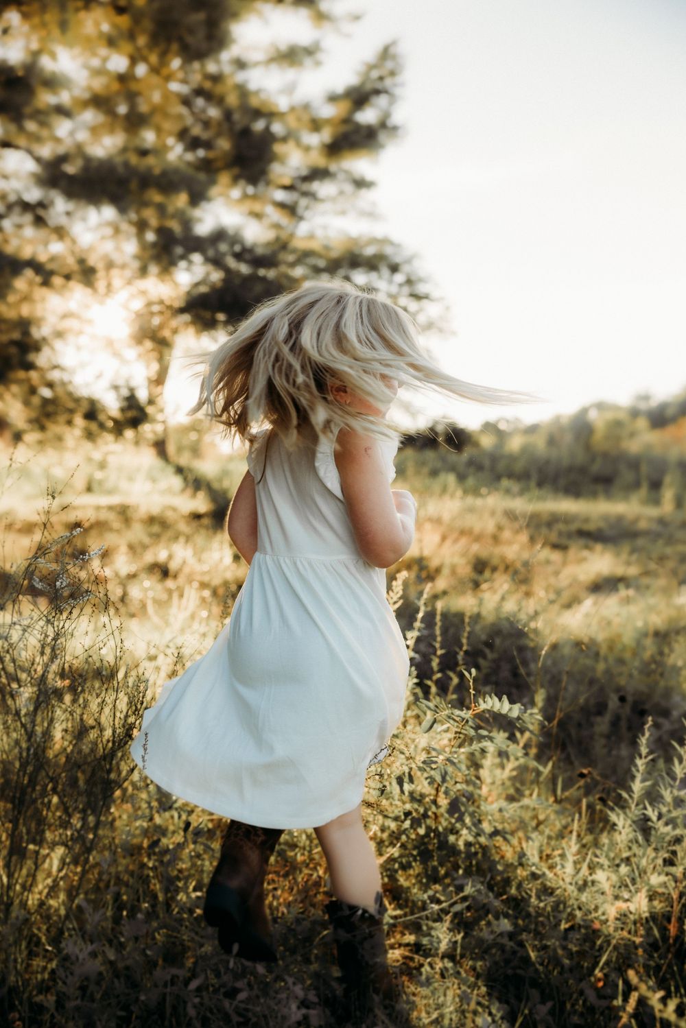 Young blonde girl in white dress and cowboy boots twirling in a sunlit field at golden hour.