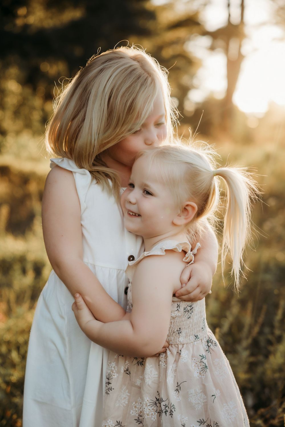 Two young blonde sisters hugging and sharing a sweet kiss in a sunlit outdoor setting.